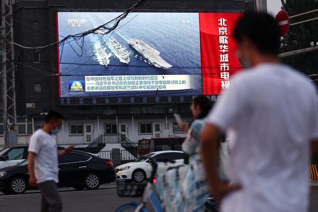 Pedestrians walk past a screen showing footage of Chinese People's Liberation Army (PLA) ships during an evening news programme, in Beijing
